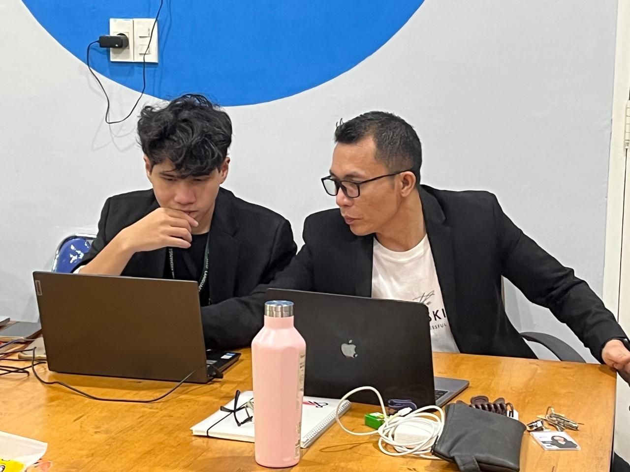 Two people in black jackets working at laptops on a wooden table with various items scattered around.
