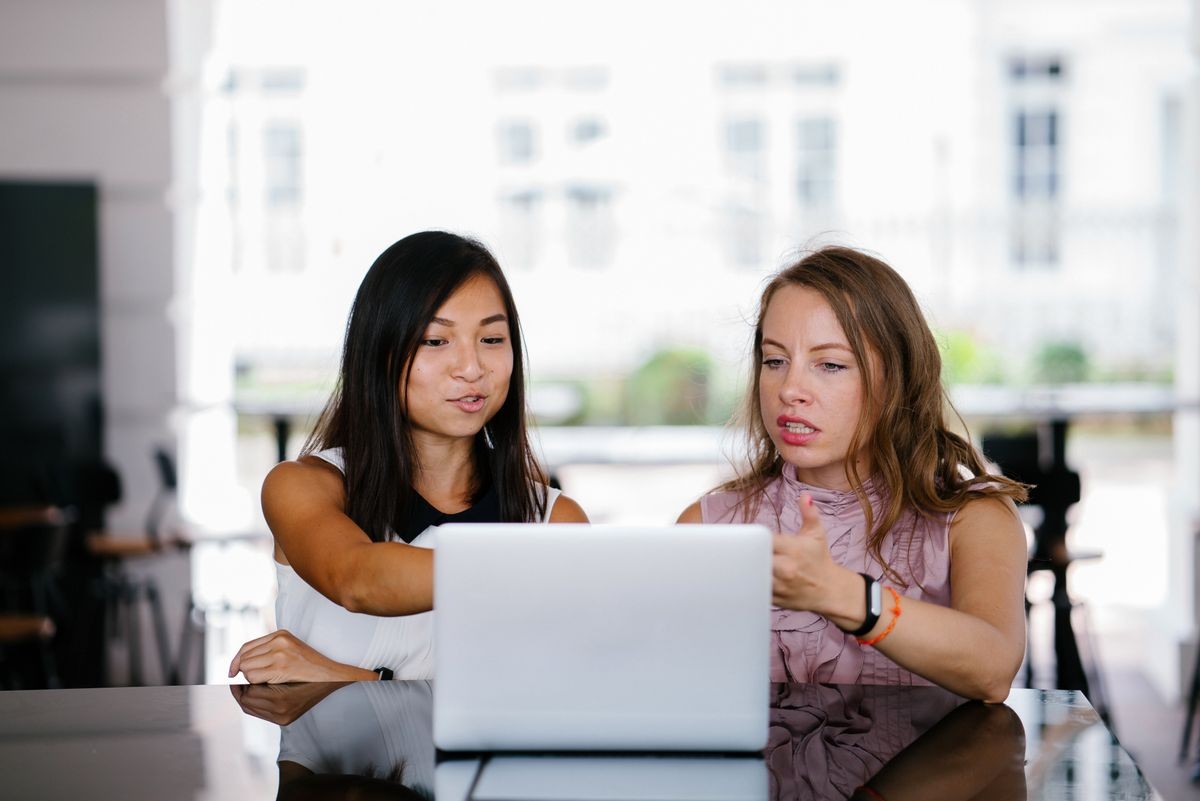 A diverse team of two coworkers (Chinese Asian woman and Caucasian white lady) working over a laptop. They are looking at the screen and discussing what is on the screen. 