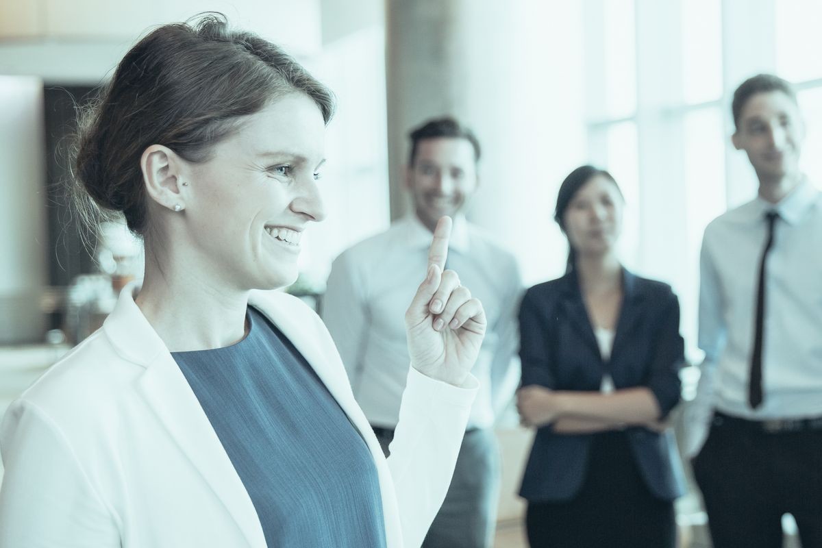 Optimistic female business leader showing index finger as symbol of idea. Cheerful executive running business, her team standing in row in background. Businesswoman concept